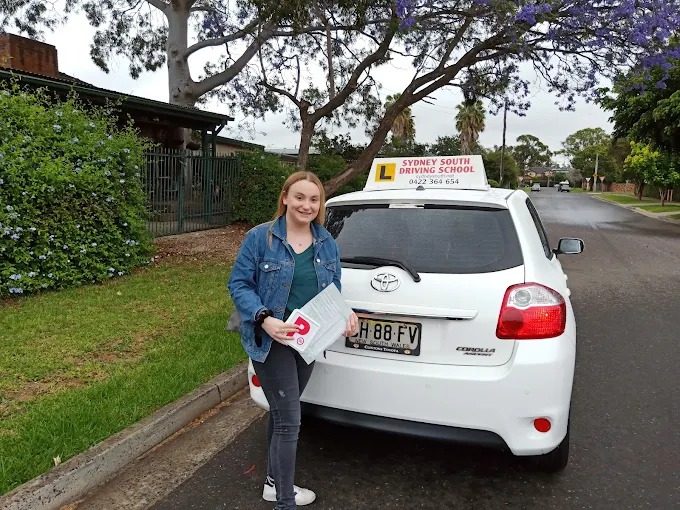 A learner driver holding a logbook beside a Sydney South Driving School car on a suburban street